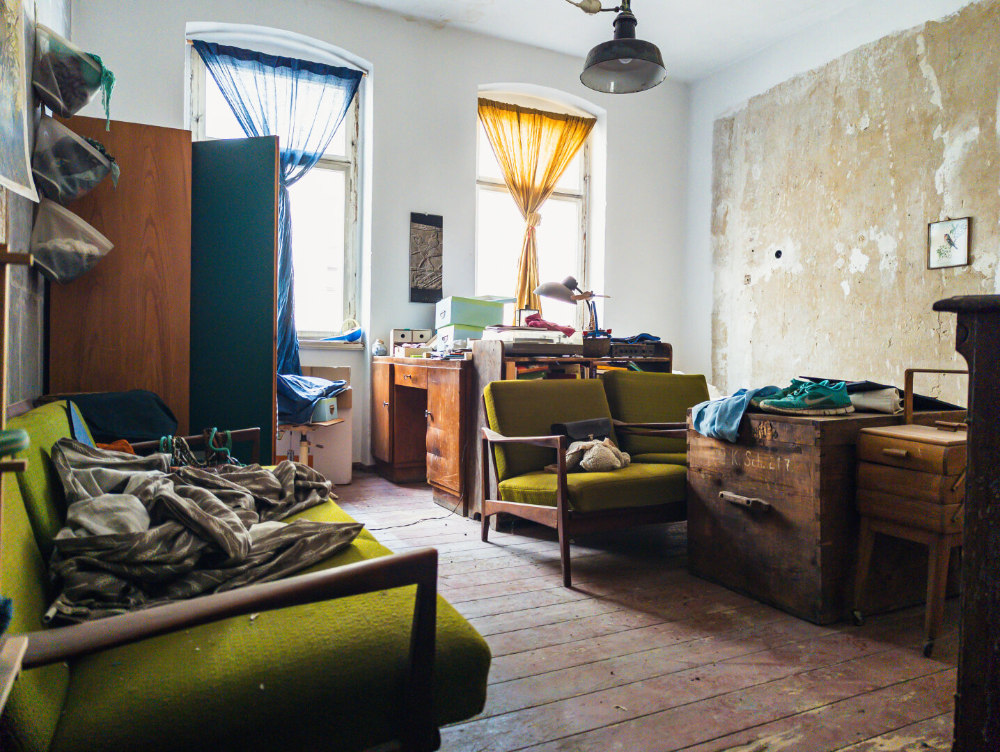An abandoned, cluttered vintage room with green furniture, draped curtains, and peeling walls.