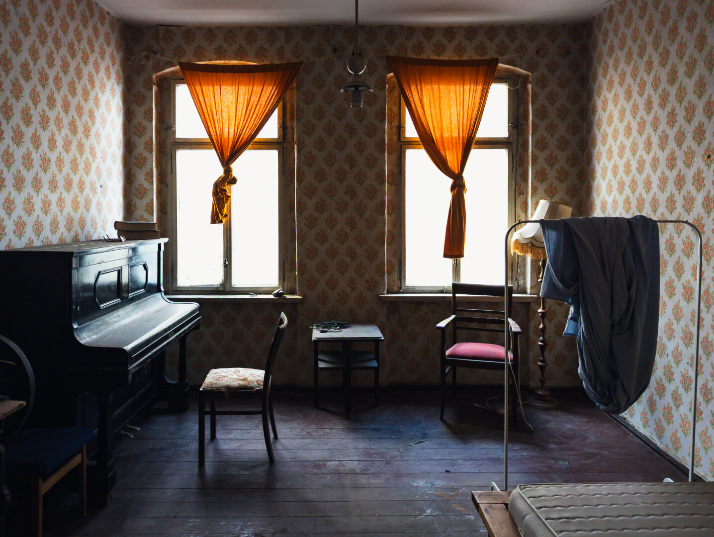 An abandoned vintage room with floral wallpaper, draped orange curtains, an upright piano, and scattered old furniture.