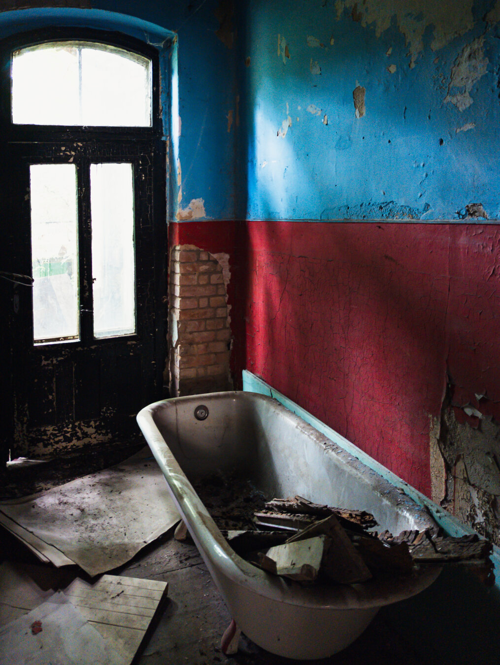 A decaying, abandoned bathroom with peeling blue and red walls, exposed brick, and a bathtub filled with debris.