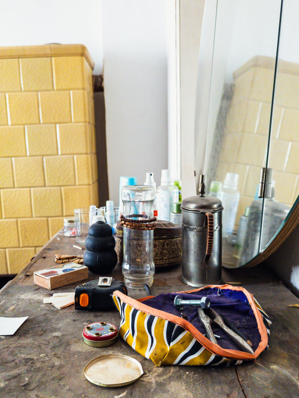 A cluttered, dusty tabletop with scattered tools, glass jars, and a yellow patterned pouch beside a yellow tiled wall and mirror.