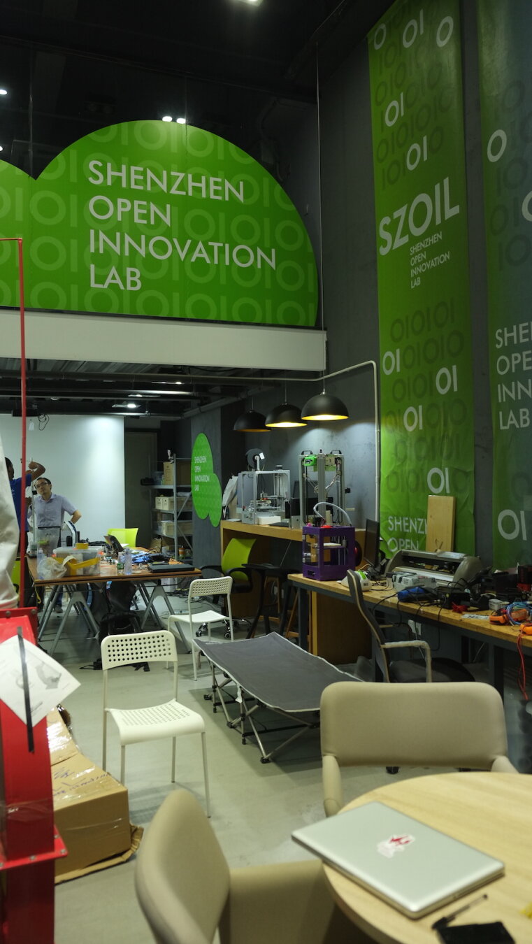Photo of a fab lab maker innovation prototyping space in Shenzhen, China. The name of the lab SHENZHEN OPEN INNOVATION LAB and SZOIL is printed on bright green banners which hang on walls and from ceilings. The space is high with a mezzanine level. On the ground floor there is a row of counters and shelves along the wall with all kinds of equipment and shelving, including 3D printers and vinyl cutters at various table heights. In the working space there are circular meeting tables and long rectangular working tables with chairs around them. A lot of maker stuff sits on shelves and tables and a foldout camp bed sits by the 3D printers. Futian District in Shenzhen, 15 August 2016. Photo Cindy Kohtala
