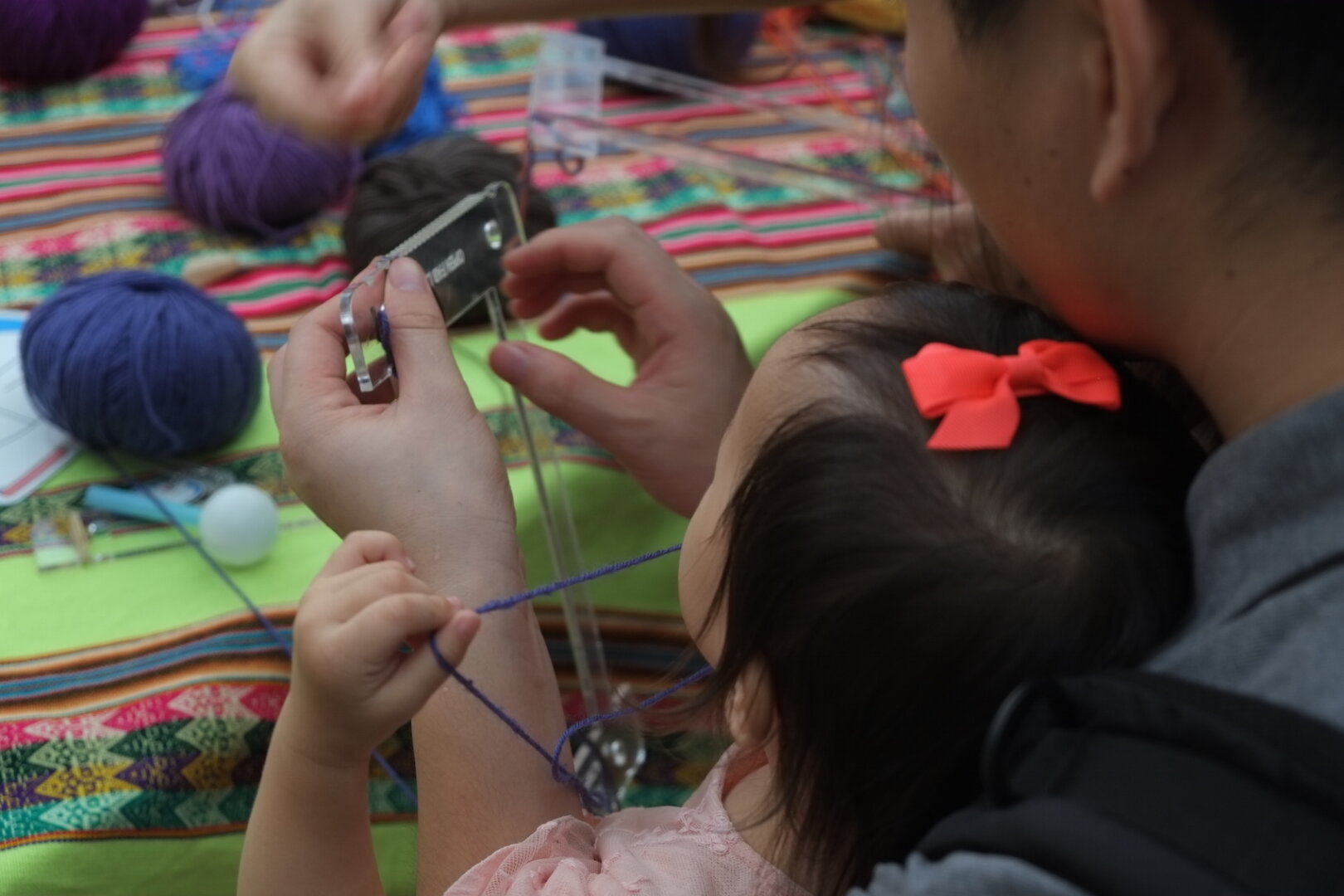 Photo of a father and daughter, a young child with a bow in her hair and sitting on his lap, from the back, focusing on their hands. They are working with a small lasercut acrylic loom. The girl is pulling on a thread of blue yarn and the father is holding the loom about to thread it. Balls of different coloured yarn are on a table in front of them. The table is covered with a brightly coloured Peruvian woven textile. FAB12: Fabricating the Future, Shenzhen, China, 13 August 2016. Photo Cindy Kohtala