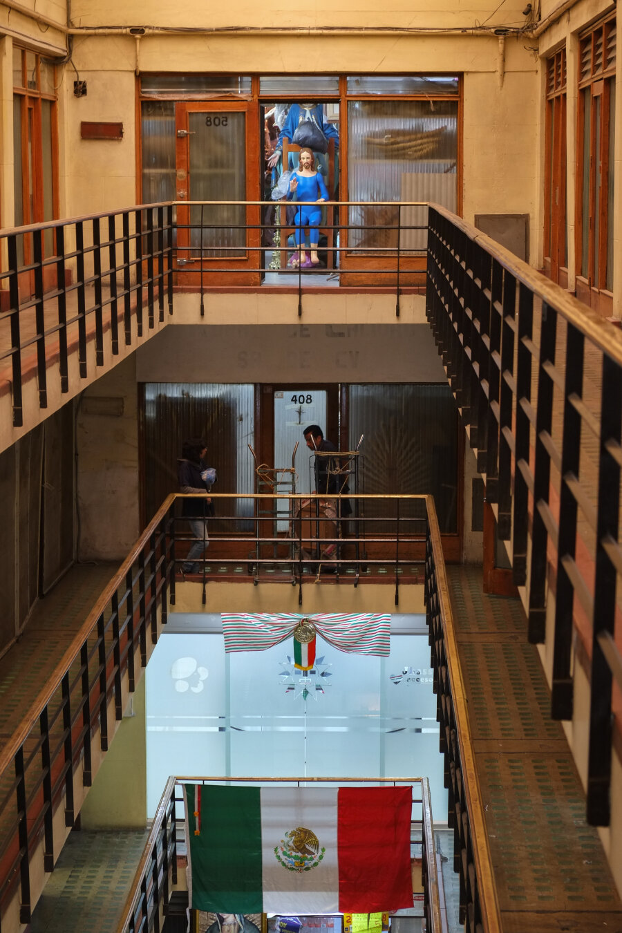 Photo inside a specialized retail building for Catholic church items. The building has a central courtyard inside that extends for the height of the building and railed mezzanine levels around the centre - from which doorways lead to businesses. Two levels down, a large Mexican flag is hanging from the railings. One level down, a delivery person is walking with goods on moving trolleys. At the same level on the other side of the building, there is an open door where a full size statue of Jesus is sitting on a large wooden chair raising one of its hands up, with some plastic wrap behind it. Other religious items are visible behind it. Right behind the main Cathedral at the Zócalo, Mexico City, 12 September 2016. Photo Cindy Kohtala