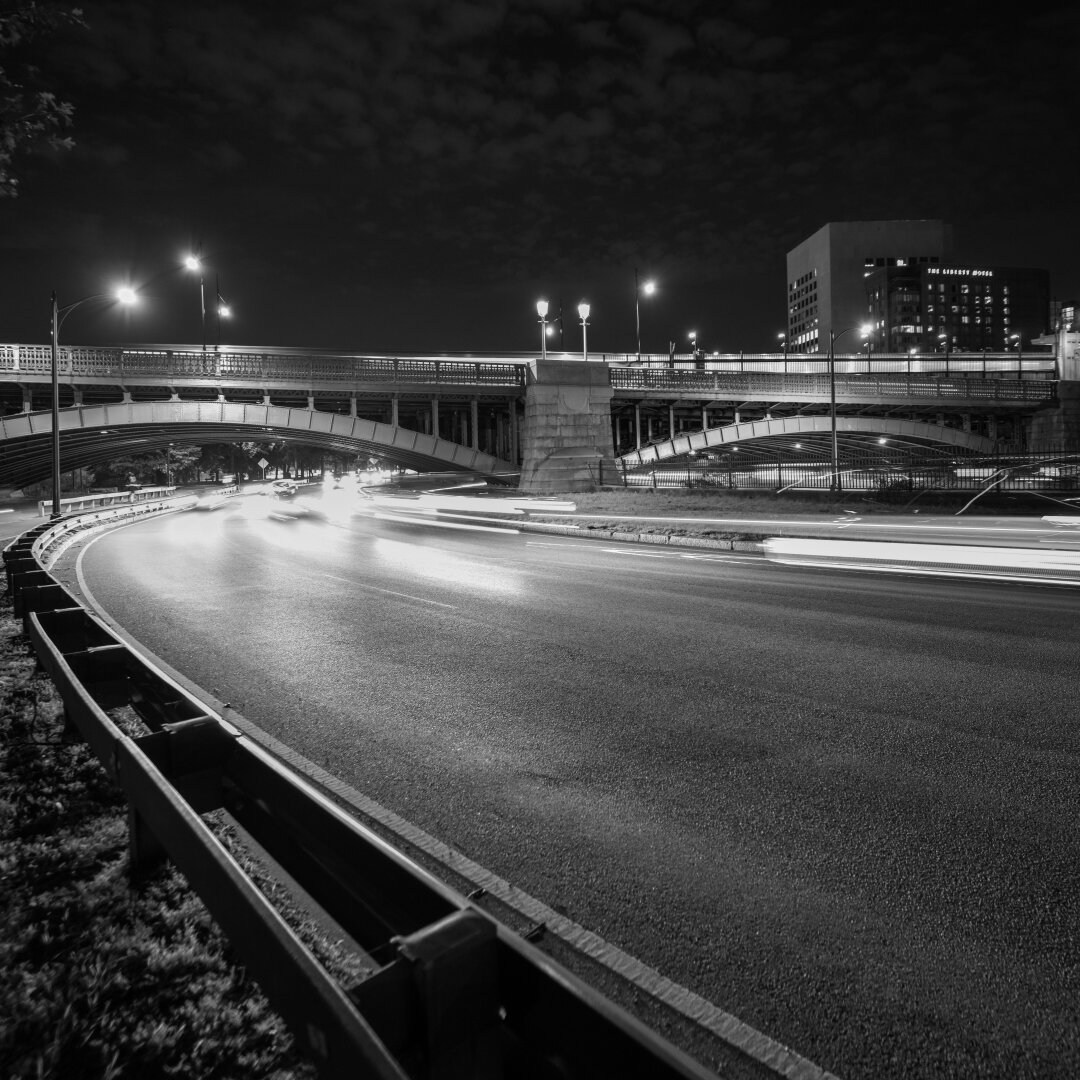 Headlights streak past a curve in the road underneath a commuter train crossing an archway bridge at night.