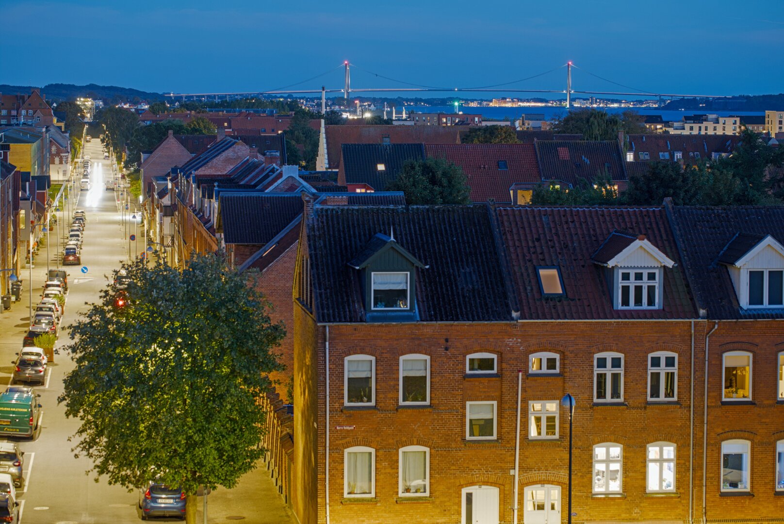 View over a small town. On the left a long, straight street leads into the distance. Cars are parked on the side and yellow streetlights illuminate it. In the foreground a large brick building with white window frames occupies the photo. In the far back you can see open water and a huge bridge over it, which leads to an island.
