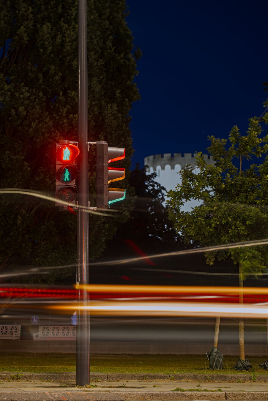 Long exposure photo of a streetlight in the dark. All three lights lit up during the exposure, so it looks like they all shine at the same time. The pedestrian lights are soldiers, both lights can be seen in the photo. The green signal is a walking soldier and the red signal is a standing soldier, both carry a large gun with them. In the background are some trees and a tall, white tower which was the old water tower of the town.