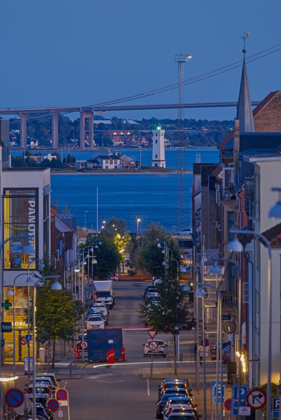 View through a long, straight street that leads to open water. There is a small white lighthouse with a green light and behind it a huge bridge and parts of the island which it leads to.