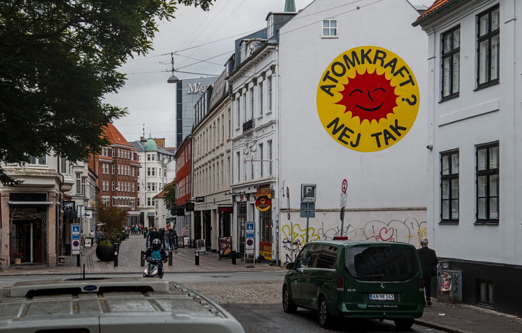 View of a street in the town of Aarhus. Some people, cars and bikes can be seen. One house wall has a large anti-atomic energy symbol painted on it with the words "Atomkraft? Nej tak" written inside it.