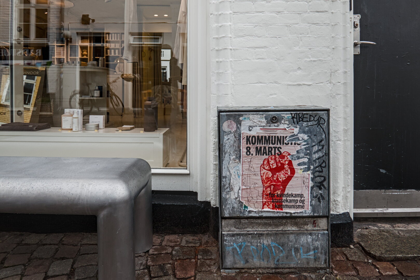 Detail view of a street, part of a round, steel bench is visible as well as an electrical box. Behind it are a door and a window from a boutique. The electric box is covered by an old poster that supports communism.