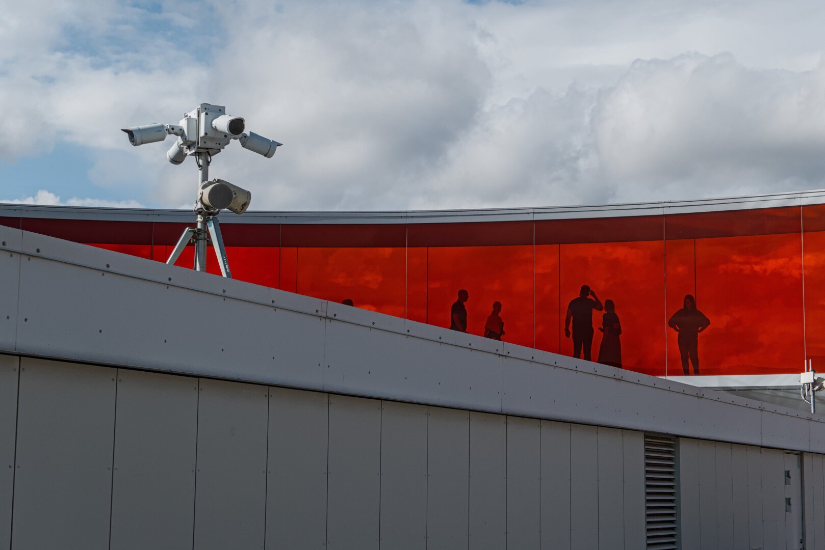 Detail view of the red part of the rainbow panorama on top of the Aros art museum in Aarhus. In front of it part of a roof can be seen with a camera pole with multiple surveillance cameras mounted on it. The silhouettes of some people can be seen inside the rainbow walk.