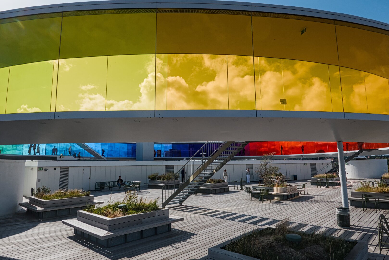 A large roof terrace with lots of places to sit and a wooden deck. The benches are mostly rectangular with some plants planted between them. A round, rainbow coloured panorama walk spans over the whole building and offers the opportunity to walk through it and see the world in different colours, because the windows are completely coloured like a rainbow.