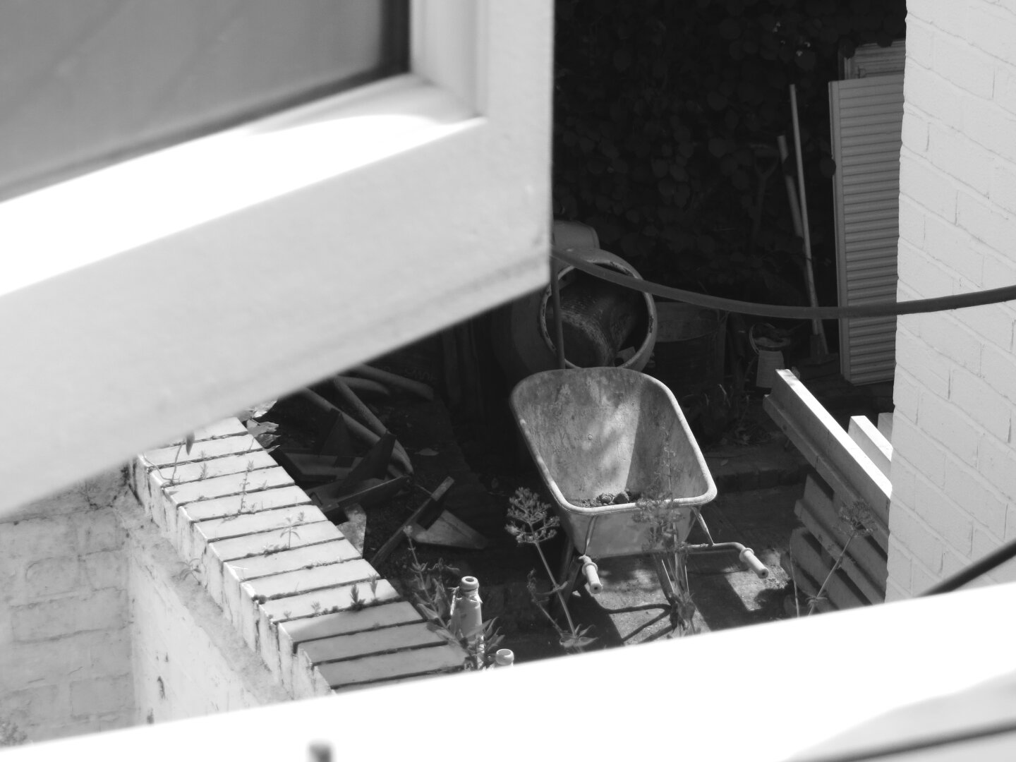 A black-and-white photograph captures a construction scene from an elevated perspective. The image is taken from above, looking down through a window. Below, a wheelbarrow sits as the subject. The ground is made of concrete slabs, some of which are slightly uneven or cracked. Various tools and equipment are scattered around the area. The scene is framed by a white wall and a window frame, adding depth and contrast to the composition.